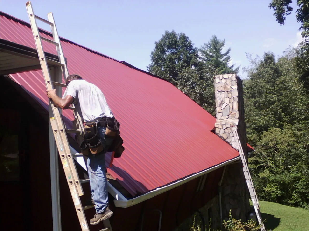 Roofer on a ladder beside a red metal roof and stone chimney — Soffit & Fascia Repair by Roofing Brothers in Coyanosa